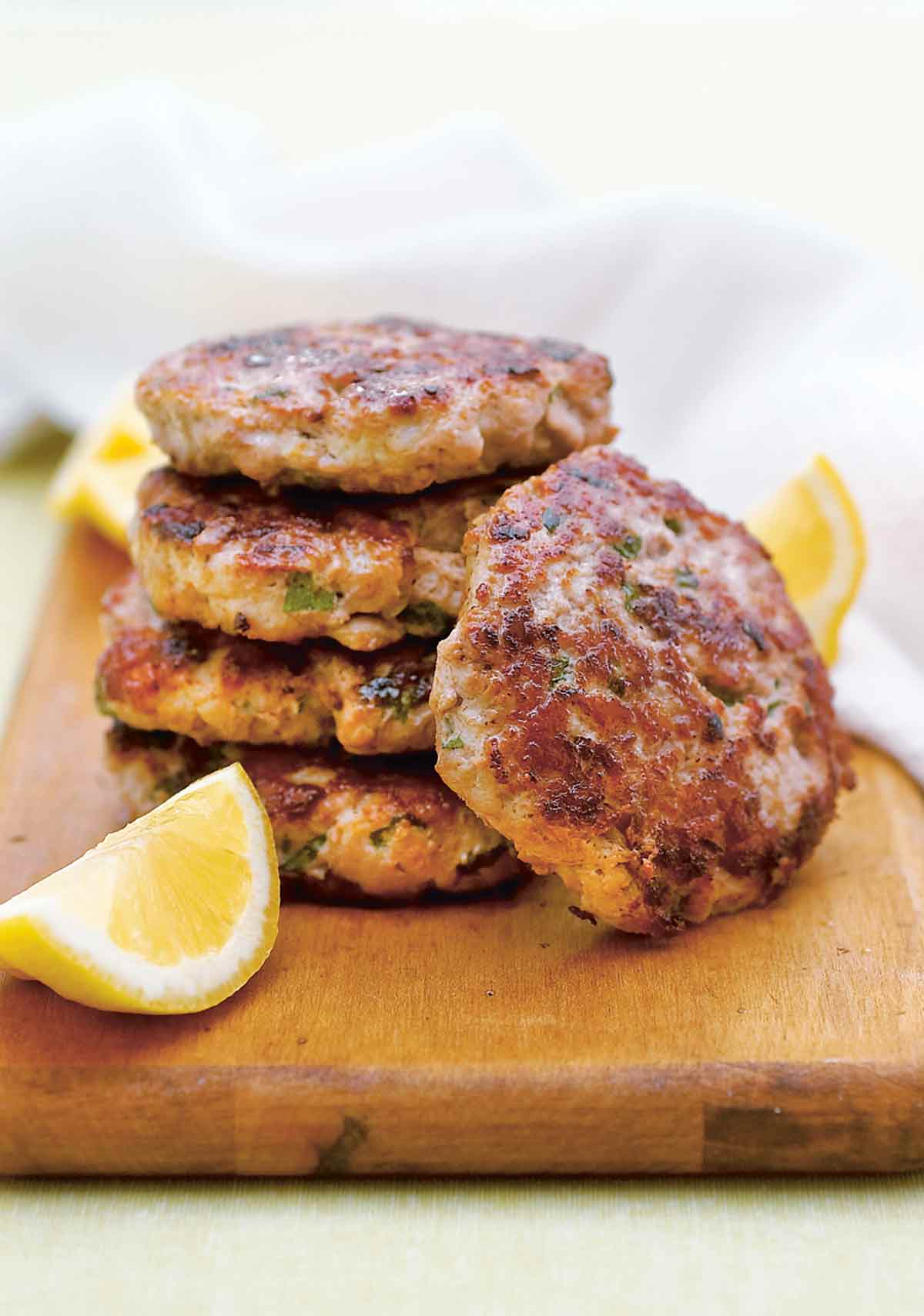 Five turkey burgers with lemon and mint stacked on a wooden board with lemon wedges.