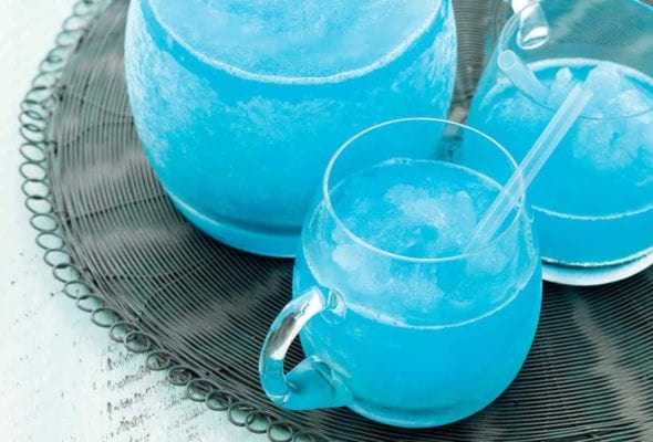 A pitcher and two glasses of blue margarita on a grey woven placemat.