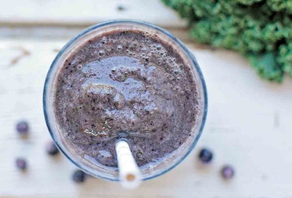An overhead view of a glass of blueberry kale smoothie with a straw and some blueberries and kale scattered around the glass.