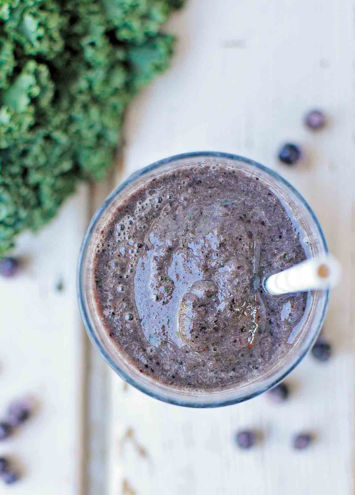 An overhead view of a glass of blueberry kale smoothie with a straw and some blueberries and kale scattered around the glass.