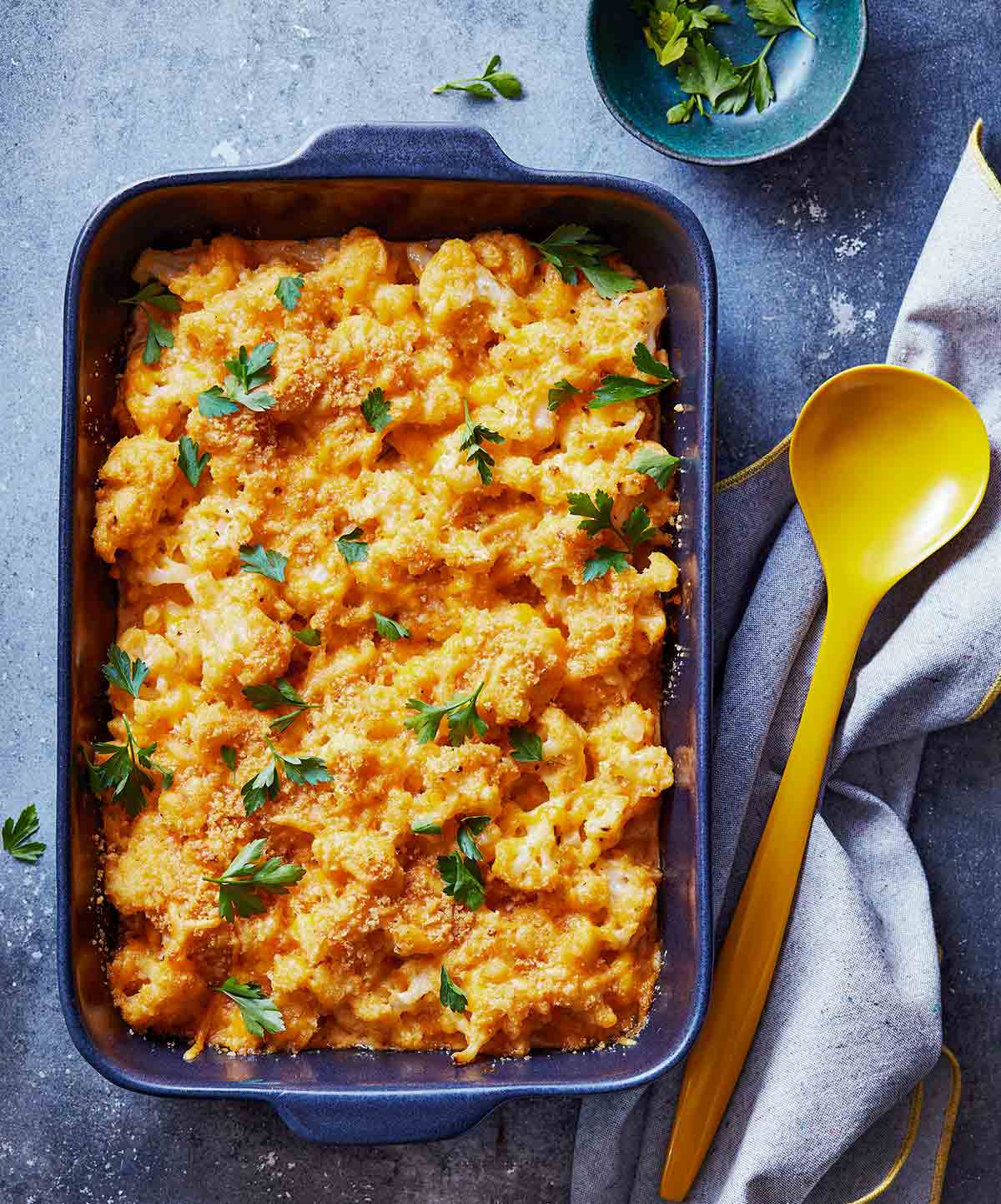 A blue casserole dish filled with cauliflower macaroni & cheese with a yellow ladle and a bowl of herbs and napkin on the side.