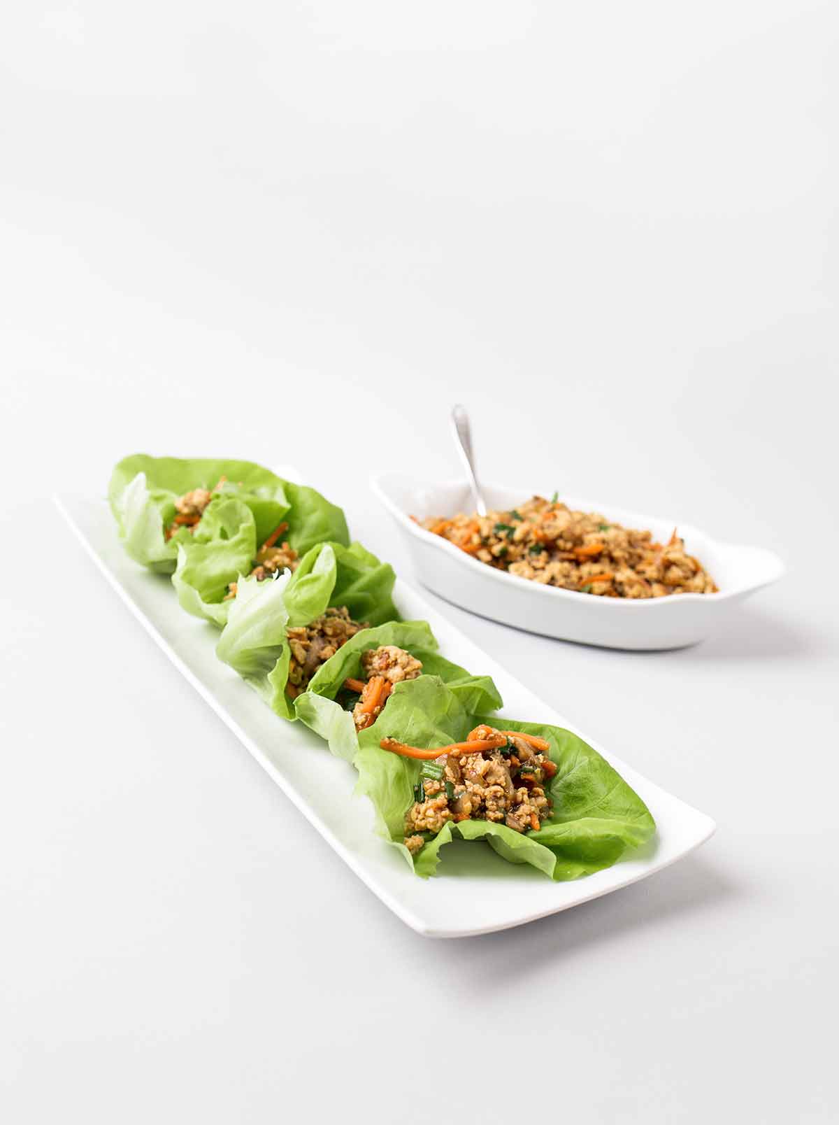 A rectangular plate with six Chinese chicken lettuce wraps and a bowl of extra filling beside it.