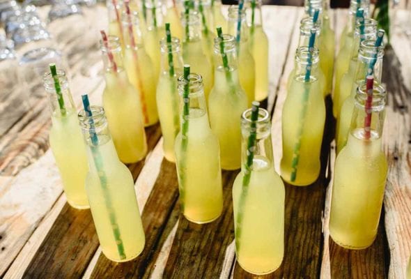 A glass bottle filled with homemade lemonade syrup, a bowl of lemons, and two glasses on a wooden table.