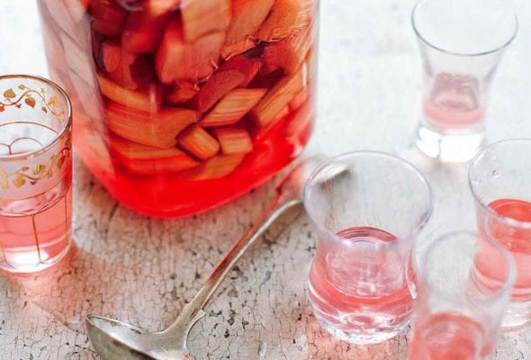 A canning jar filled with rhubarb vodka and pieces of rhubarb with several crystal glasses partially filled with rhubarb vodka beside it.