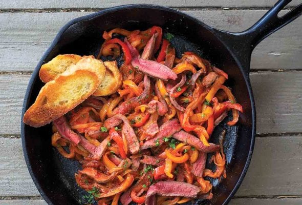 A cast-iron pan filled with skillet steak peperonata and two slices of toasted baguette.