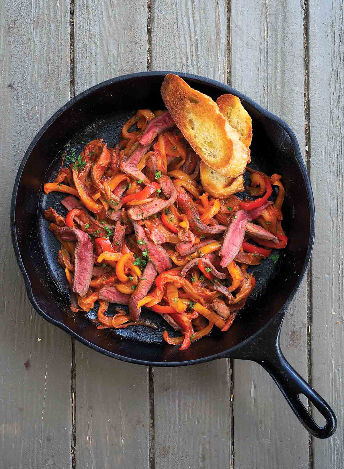 A cast-iron pan filled with skillet steak peperonata and two slices of toasted baguette.