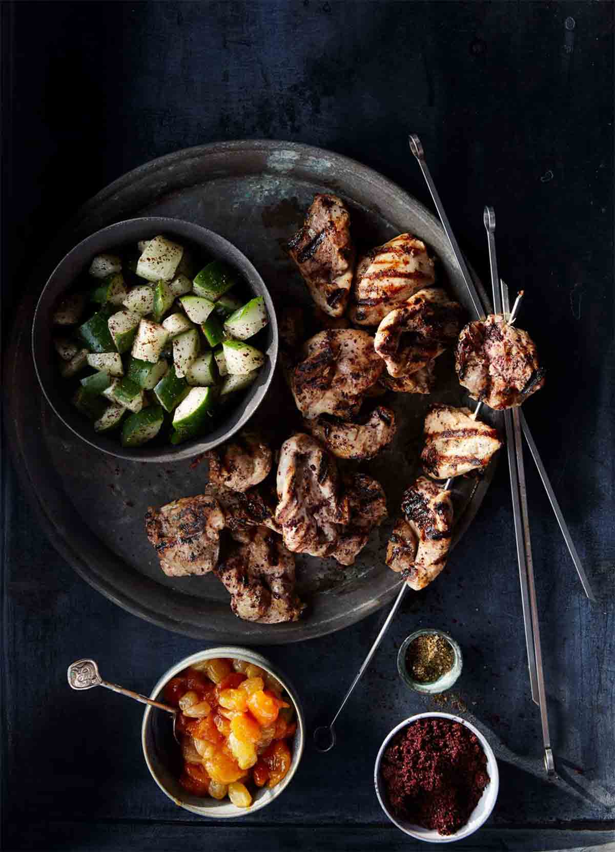 A metal plate with chunks of cooked turkey, a turkey kebab, and a dish of cucumber salad.