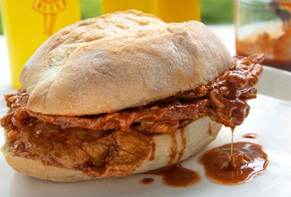 A bifana, a Portuguese pork sandwich on a white plate with a bottle of mustard in the background.