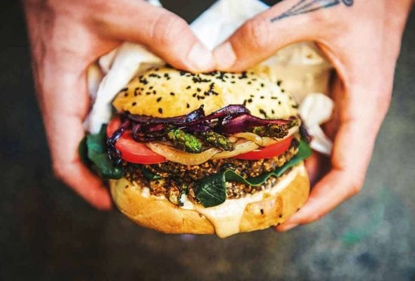 A person holding a black bean burger filled with spinach, tomato, onion, and asparagus.