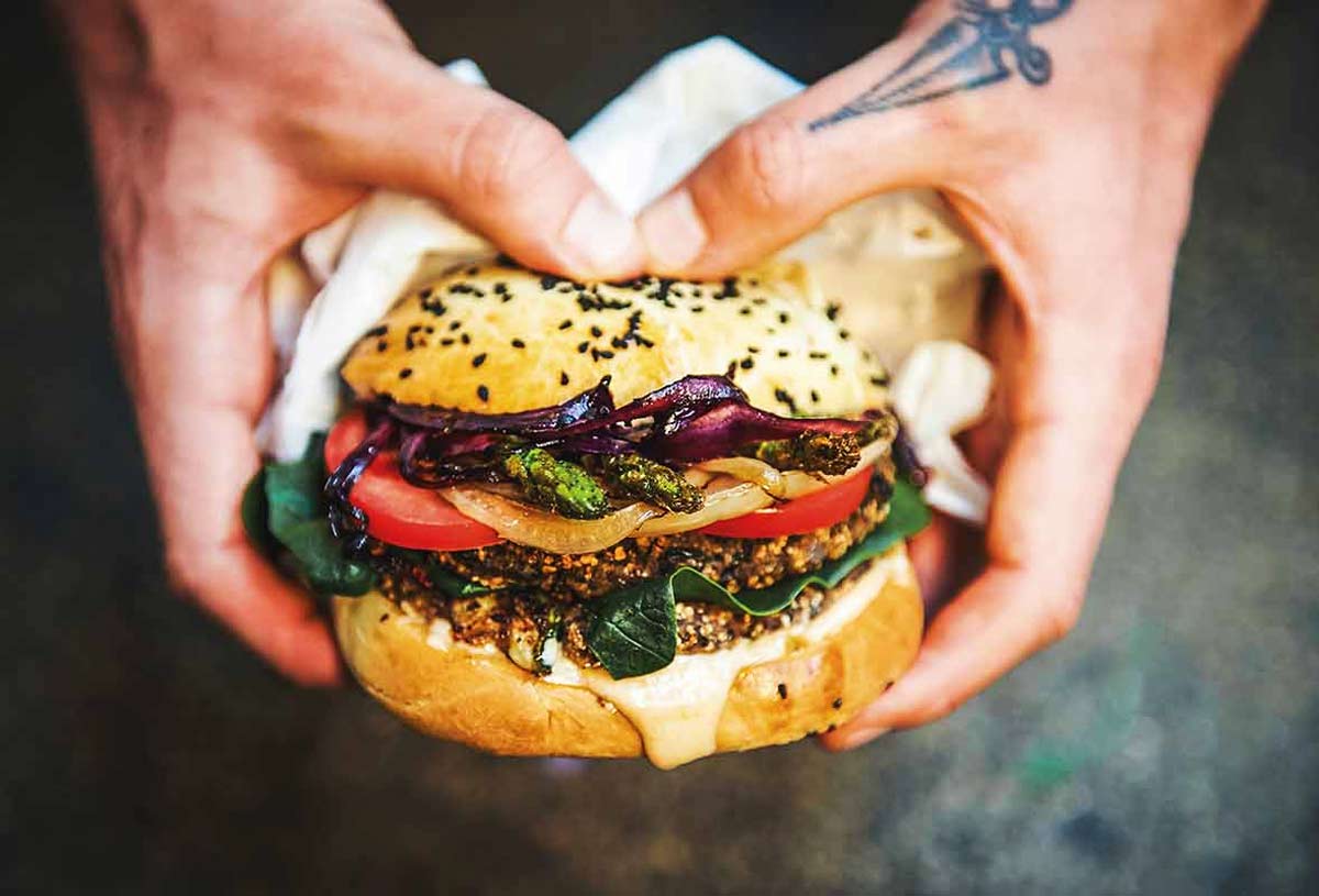 A person holding a black bean burger filled with spinach, tomato, onion, and asparagus.