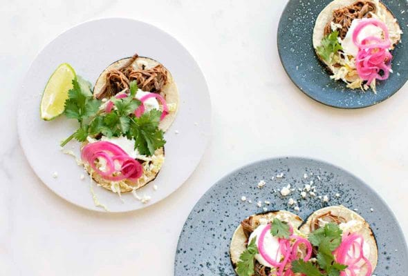 three plates filled with beef brisket tacos--braised beef brisket, Cotija cheese, napa cabbage, sour cream, red onions, and cilantro on charred corn tortillas