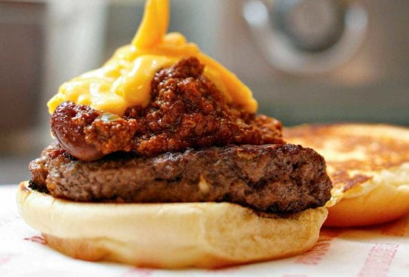A chili cheddar burger on a bun half with cheese sauce being poured over it.