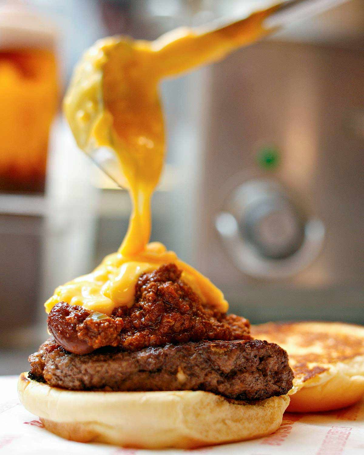 A chili cheddar burger on a bun half with cheese sauce being poured over it.