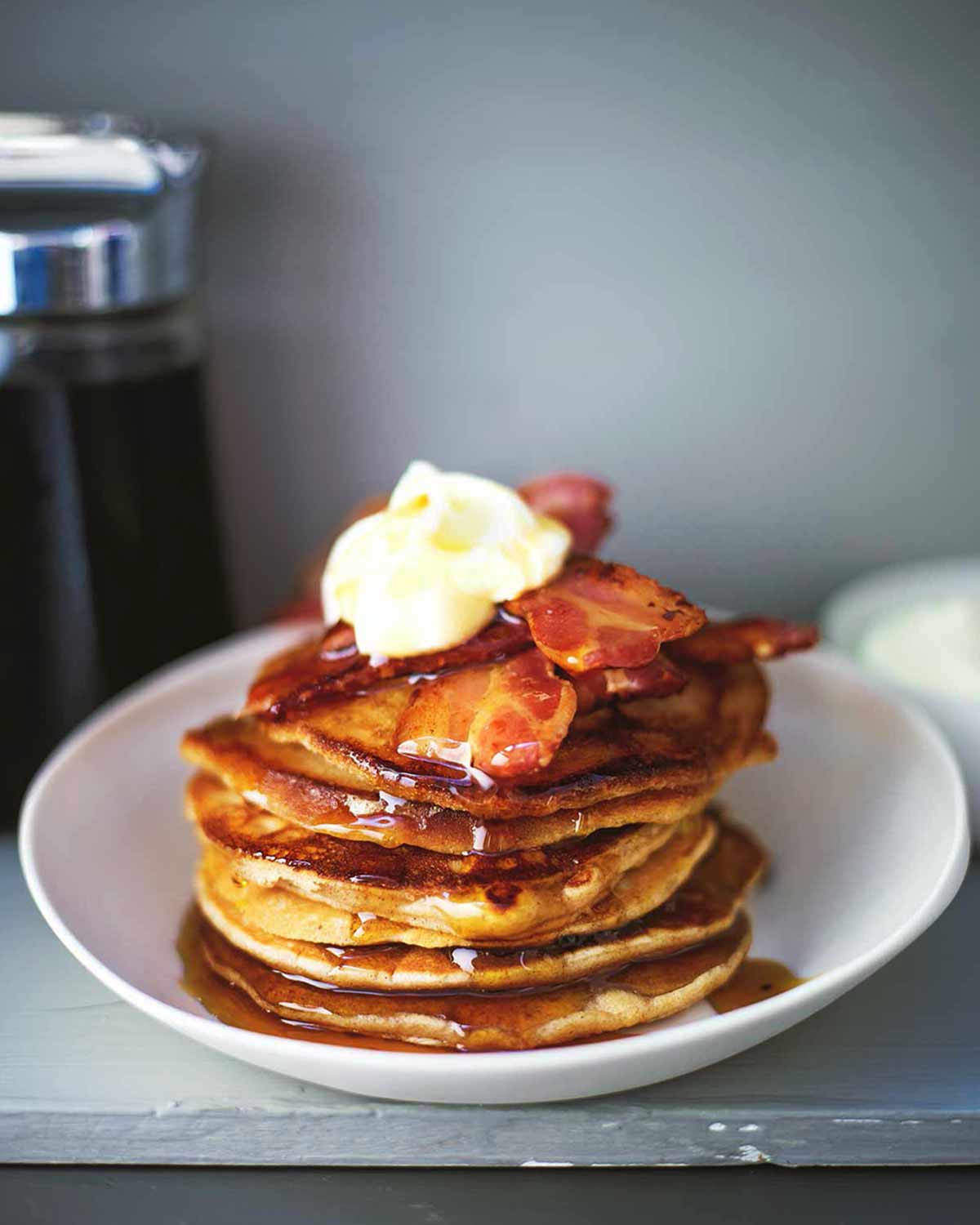 A stack of easy gingerbread pancakes topped with bacon, maple syrup, and creme fraiche in a white bowl.