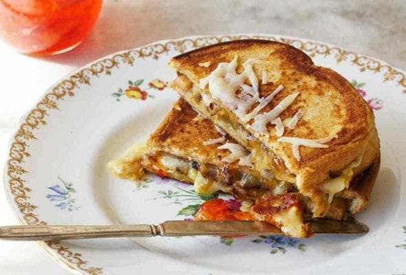 A grilled cheese sandwich with peppers is cut in half and stacked on a china plate with a jar of pepper jelly on the side