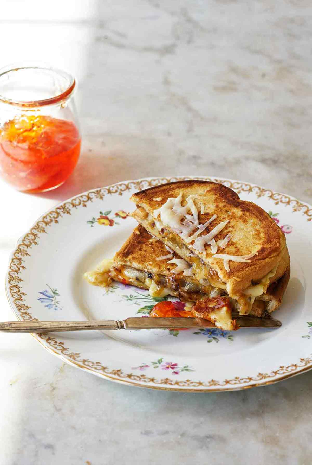 A grilled cheese sandwich with peppers is cut in half and stacked on a china plate with a jar of pepper jelly on the side