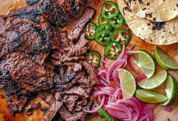 Cutting board with grilled skirt steak tacos, sliced jalepenos, tortillas, lime wedges, pickled onions, cilantro