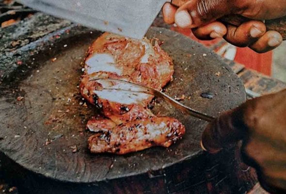 A sliced jerk chicken breast on a round cutting board, with a man's hand holding a cleaver above it
