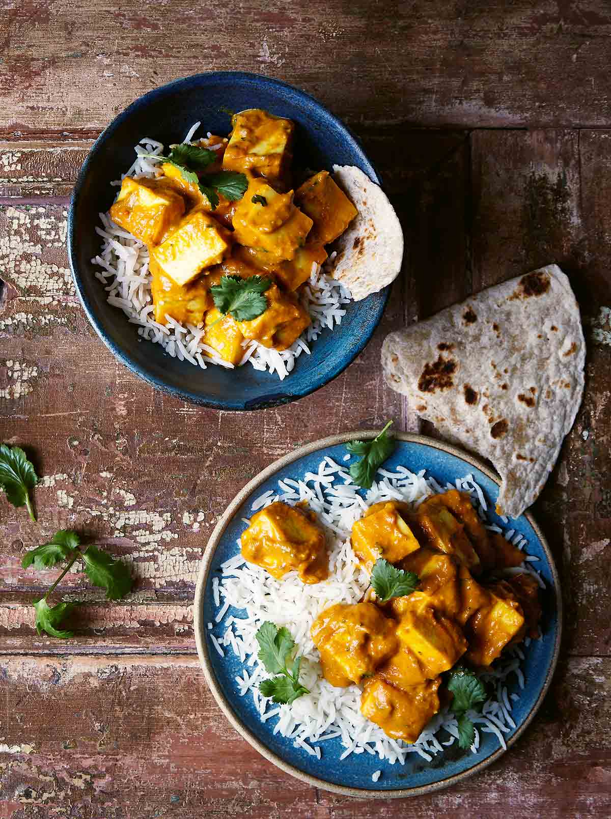 A blue bowl and a blue plate both topped with rice, naan, and masala paneer, and garnished with cilantro.
