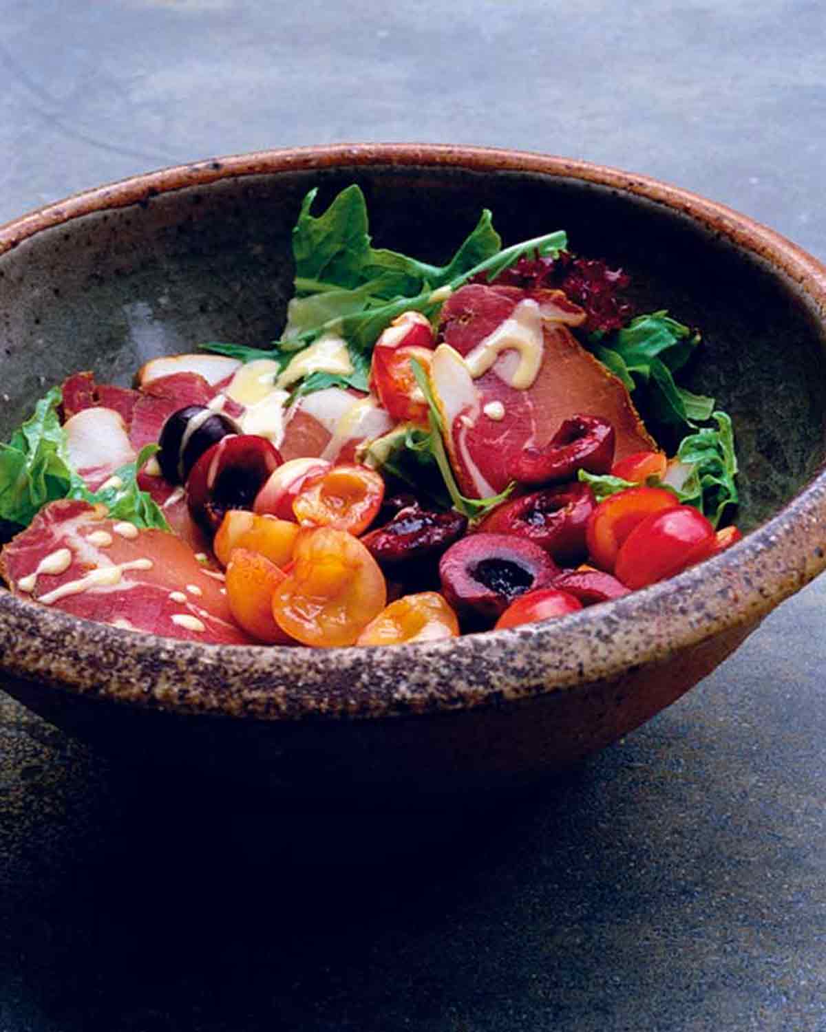 A ceramic bowl filled with mixed green salad with Prosciutto and cherries and drizzled with creamy Dijon dressing.