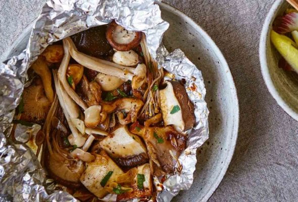 Two pouches of baked mushrooms in foil packets with a bowl of greens and another bowl of quinoa on the side.