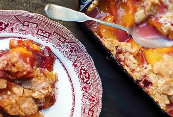 A spoon resting in a dish of cornmeal drop-biscuit peach cobbler.