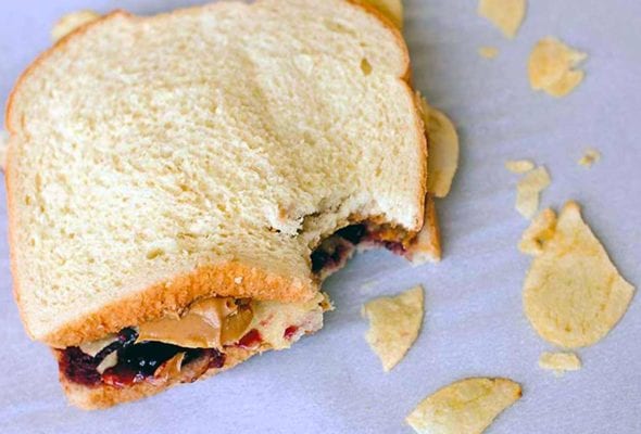 A peanut butter and jelly sandwich with potato chips with one bite gone and some chips scattered around it.