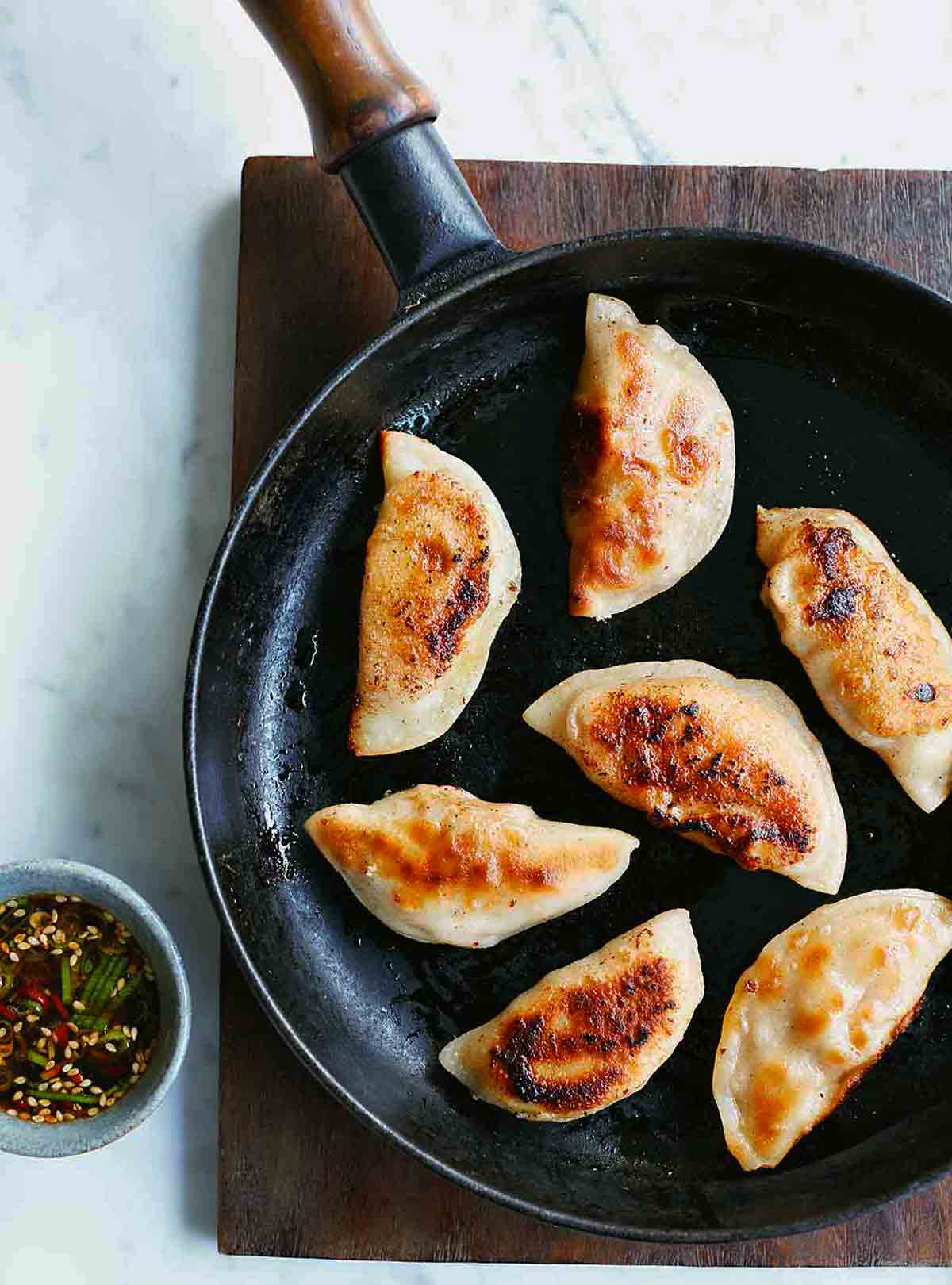 A black skillet with seven pork dumplings in it on a wooden surface and a cup of dipping sauce beside it.