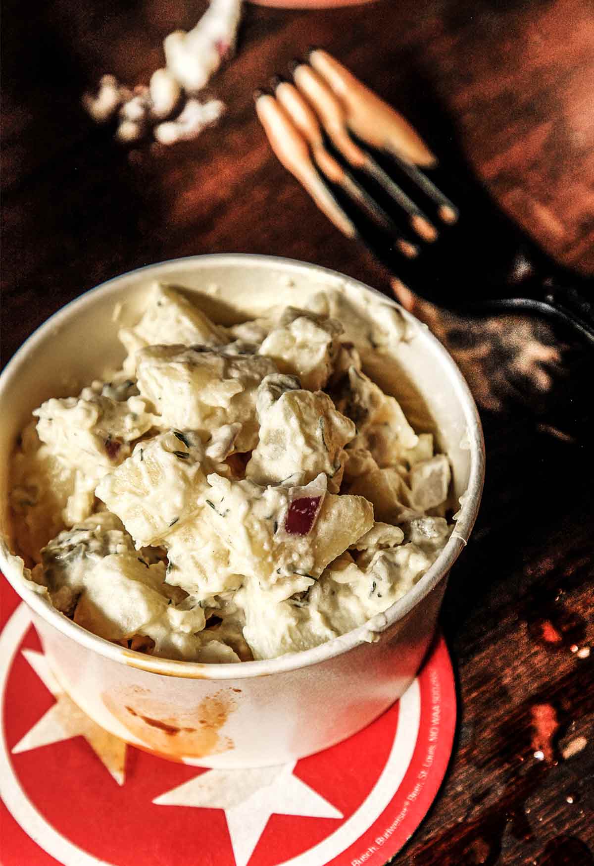 A paper bowl filled with potato salad with apples on a red and white coaster with a plastic fork beside it.