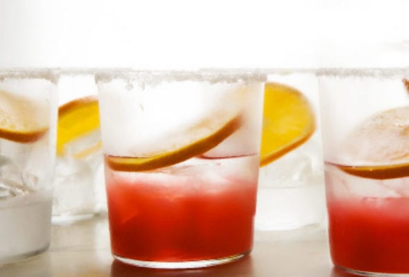 Two glasses filled with rhubarb and tequila cocktail with an orange wheel with three more glasses ready to be filled.