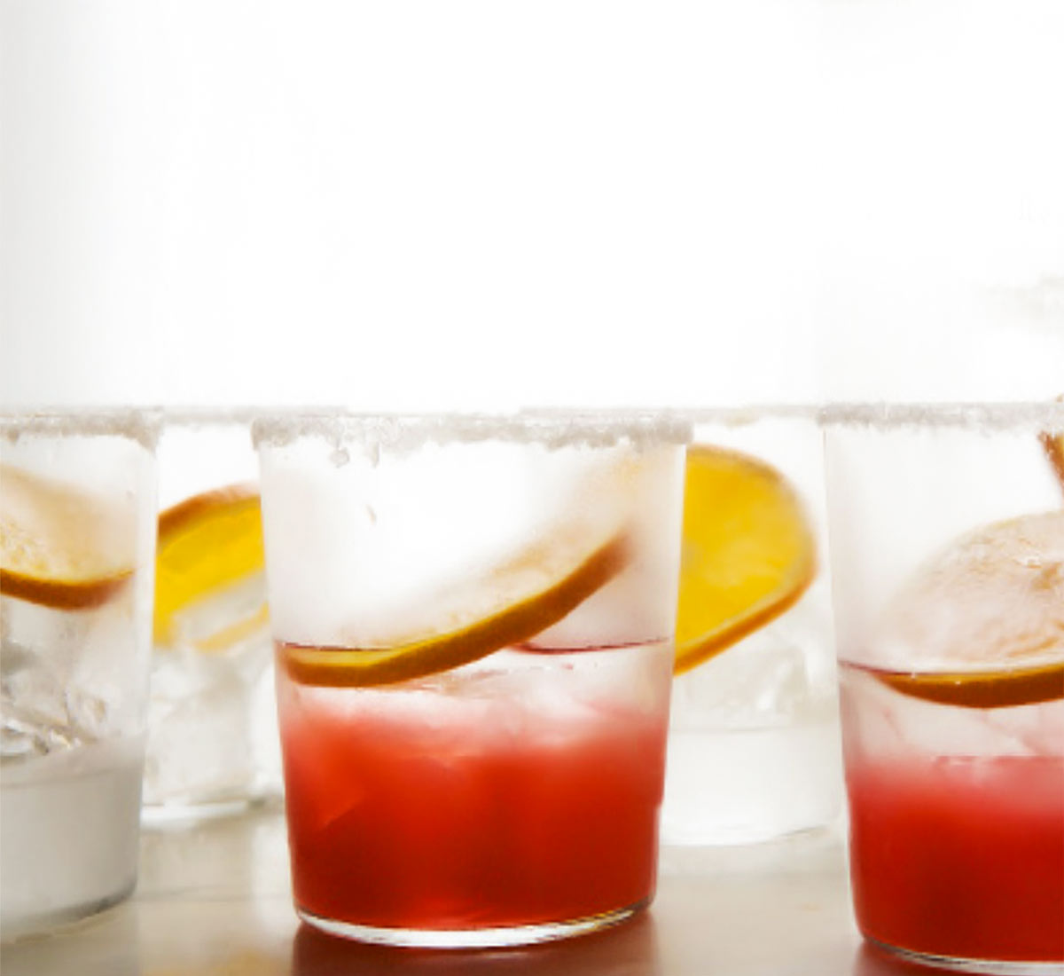 Two glasses filled with rhubarb and tequila cocktail with an orange wheel with three more glasses ready to be filled.