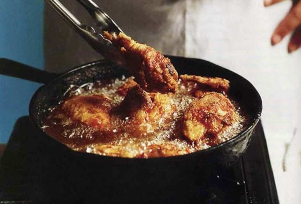 A man holding tongs in front of a skillet making southern pan-fried chicken, which is bubbling in the pan