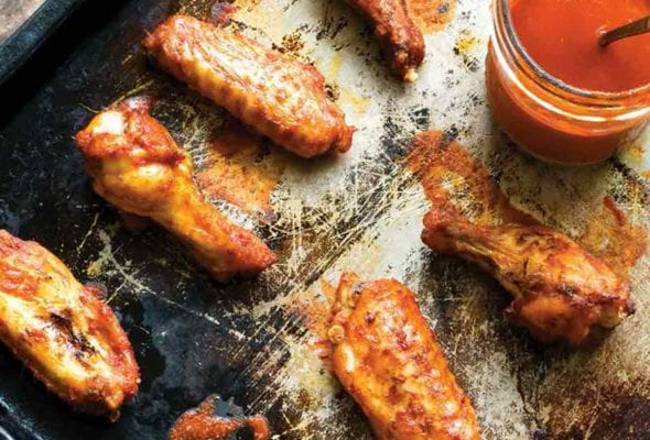 A baking sheet with 6 spicy baked chicken wings glazed with hot sauce, a jar of sauce nearby