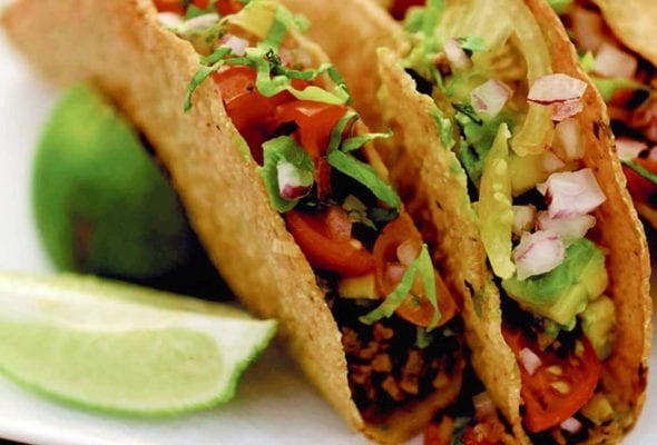 Three sweet and spicy tacos on a white plate with wedges of lime alongside.