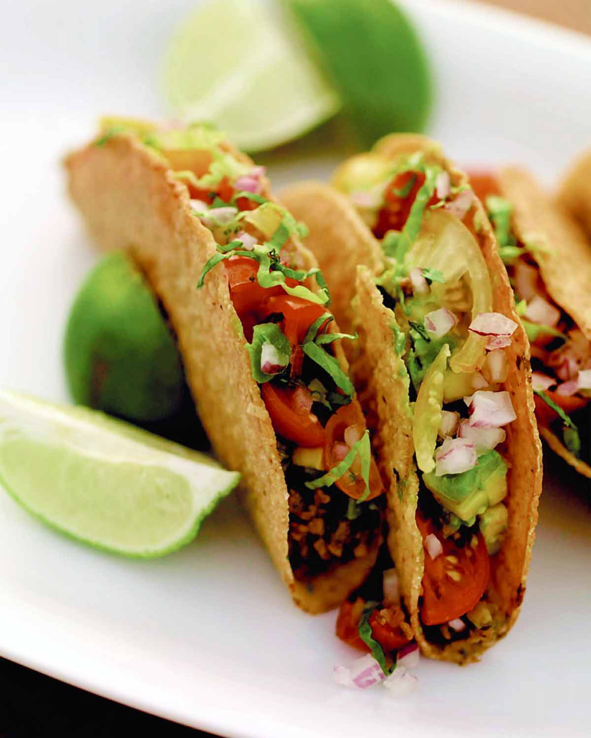 Three sweet and spicy tacos on a white plate with wedges of lime alongside.