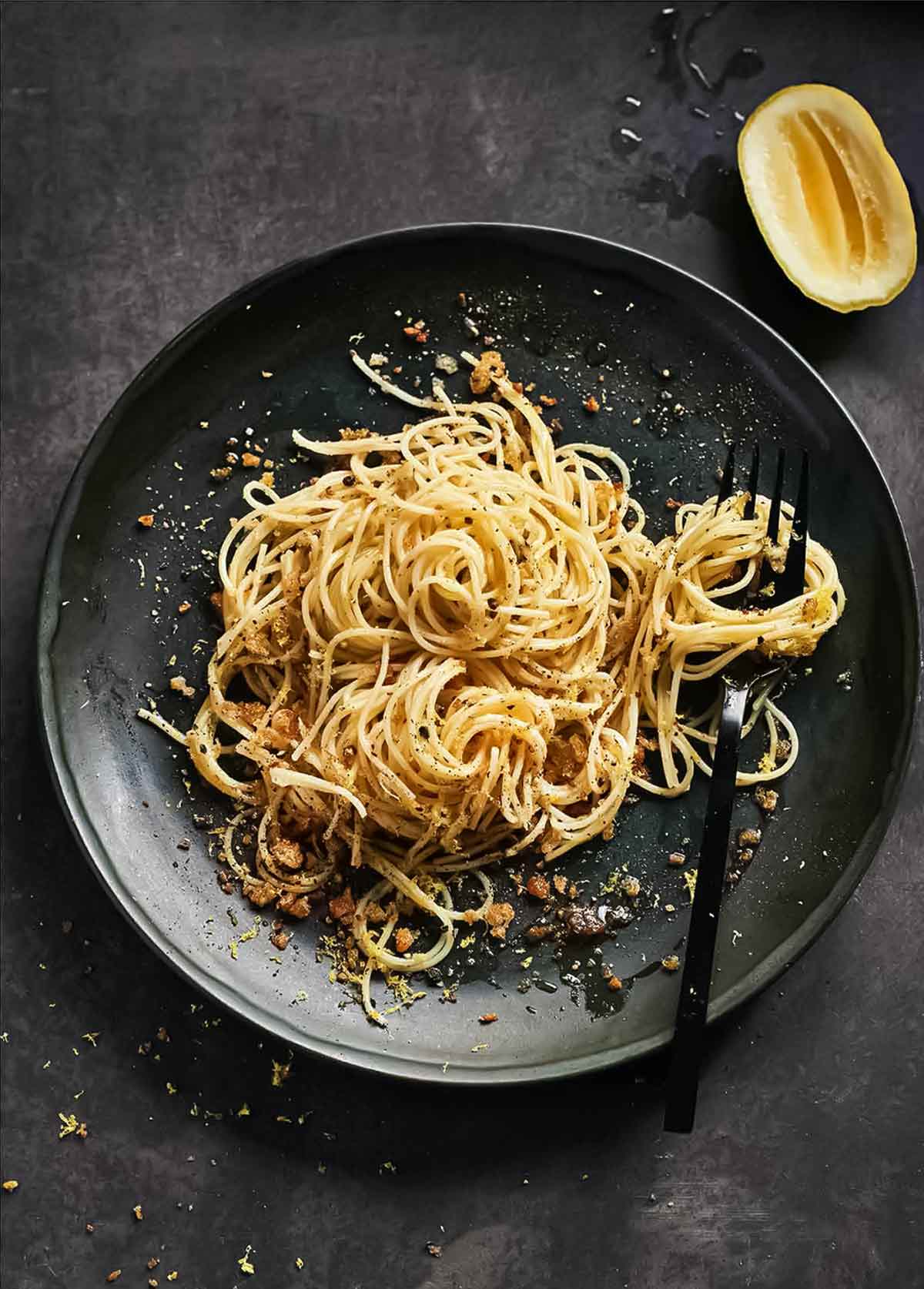 A black plate topped with angel hair pasta with lemon bread crumbs and a fork and a squeezed half lemon rests beside the plate.