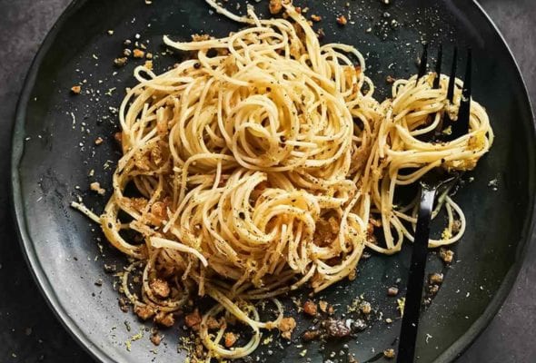 A black plate topped with angel hair pasta with lemon bread crumbs and a fork and a squeezed half lemon rests beside the plate.