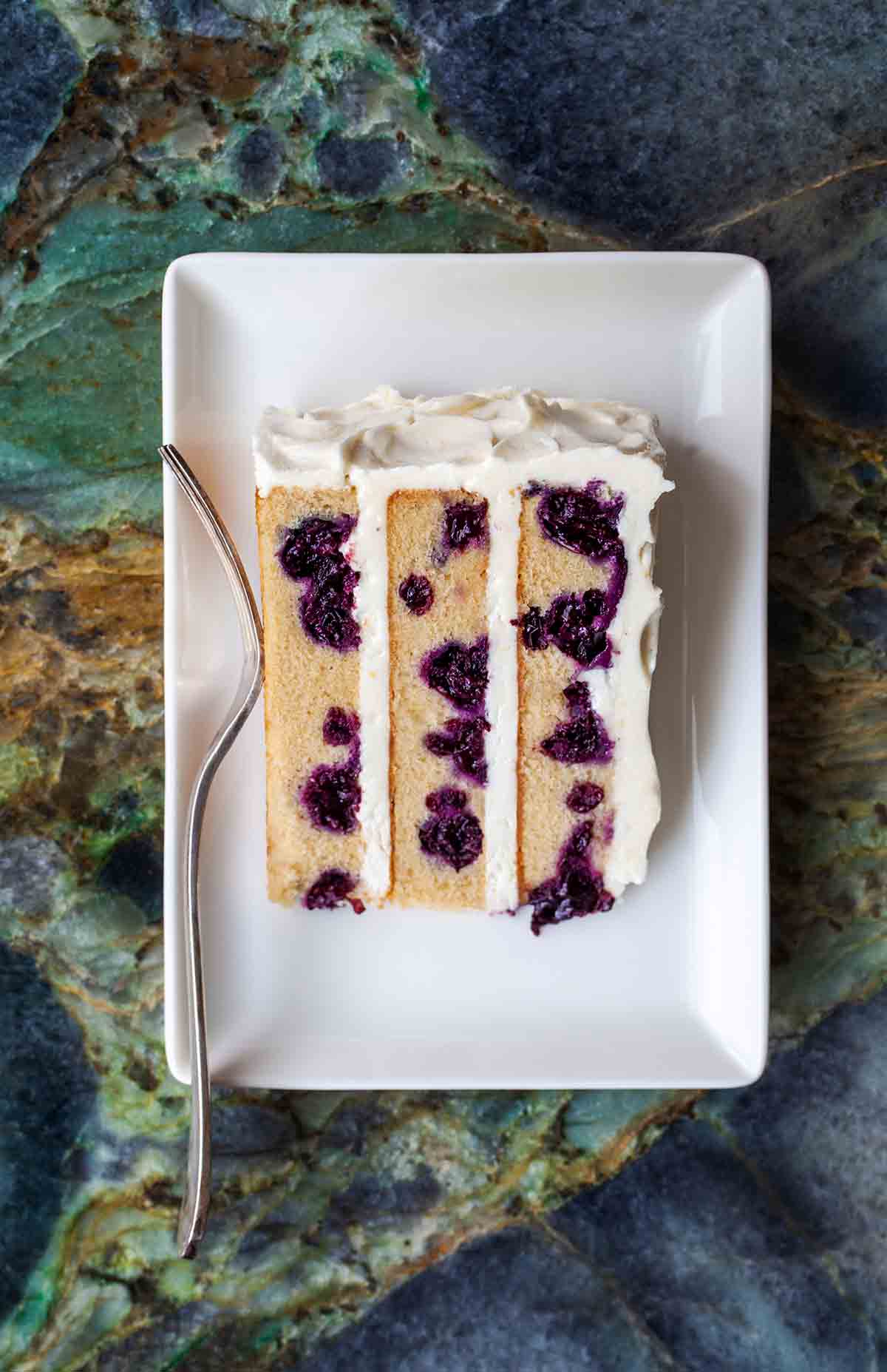 A white plate with a slice of blueberry-lemon layer cake