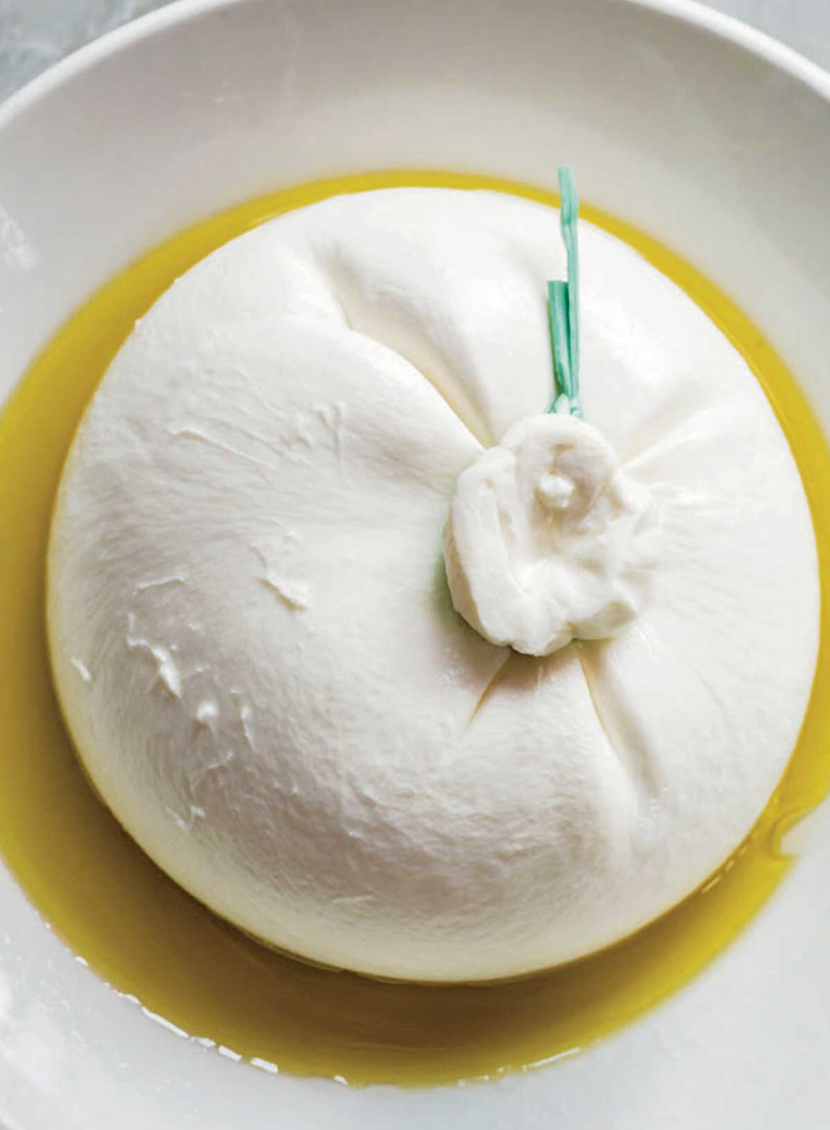 A ball of burrata for burrata with grilled bread sitting in a few tablespoons of oil in a white bowl.