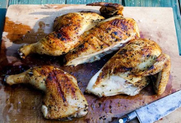 A wooden cutting board with a carved grilled Greek chicken and a knife resting beside it.