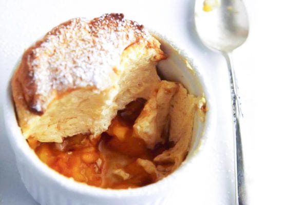 White dish of a sugar-dusted individual peach souffle; in the bottom are peach slices
