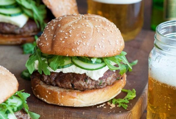 Three lamb burgers with arugula and feta on a wooden surface with two jars of beer beside them.