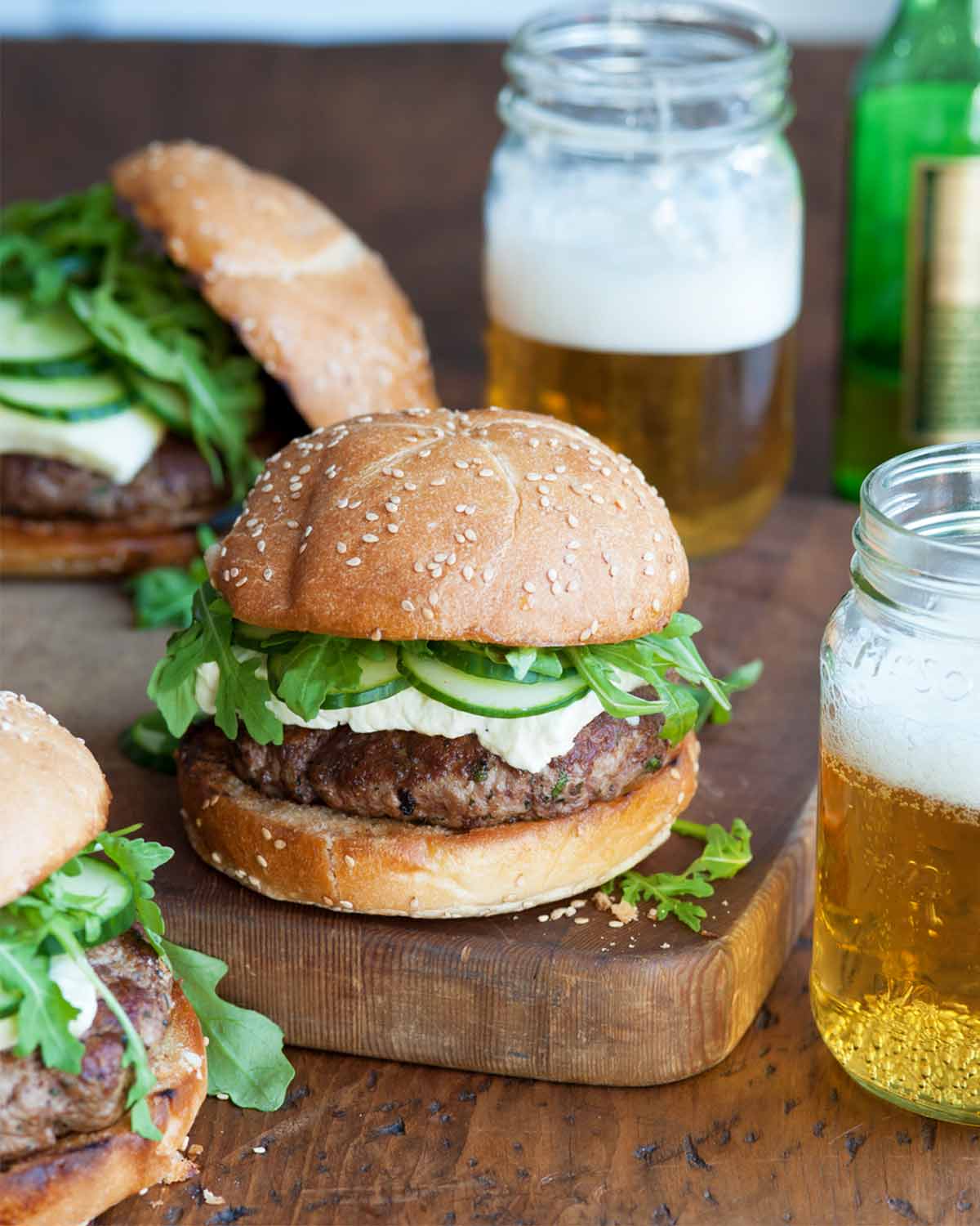 Three lamb burgers with arugula and feta on a wooden surface with two jars of beer beside them.