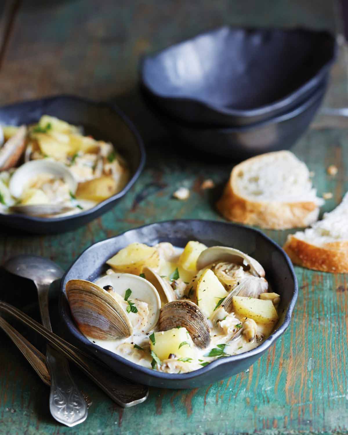 Two bowls of North Carolina clam chowder with a torn slice of bread, two empty bowls, and several spoons beside them.