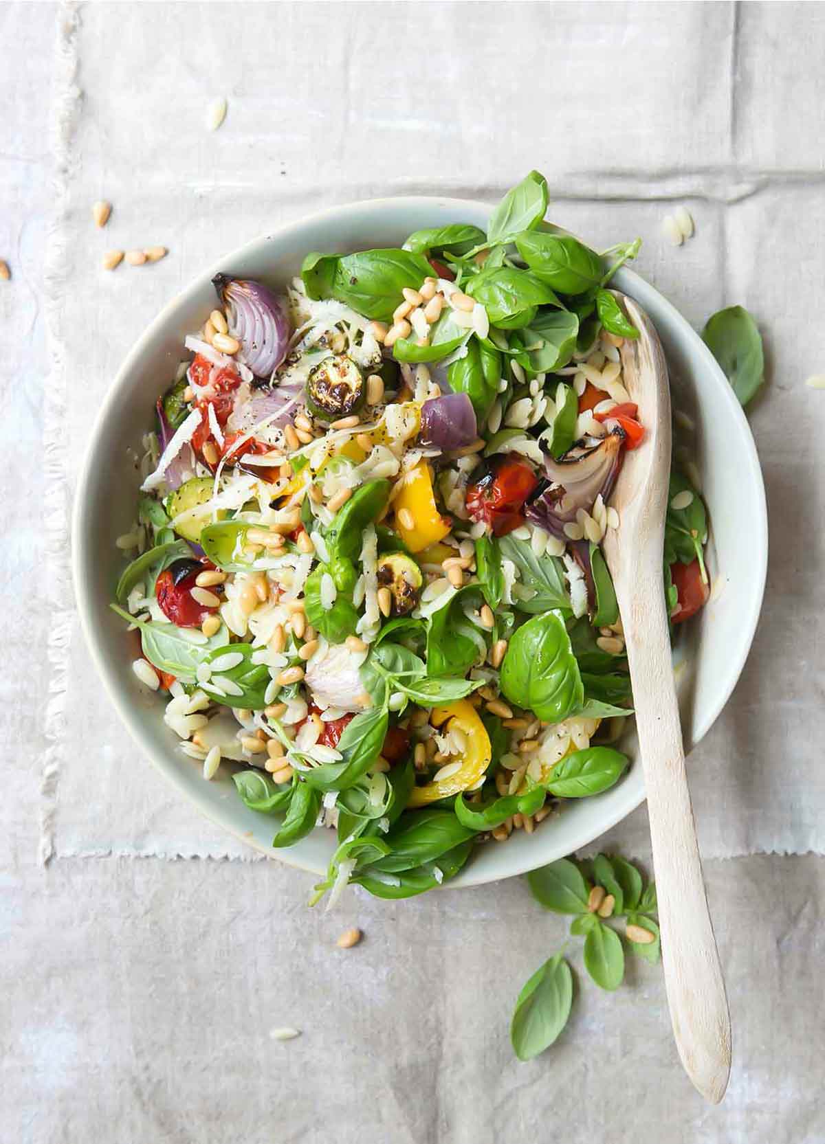 A white bowl filled with orzo with roasted vegetables, pine nuts, and fresh basil, and a wooden spoon.