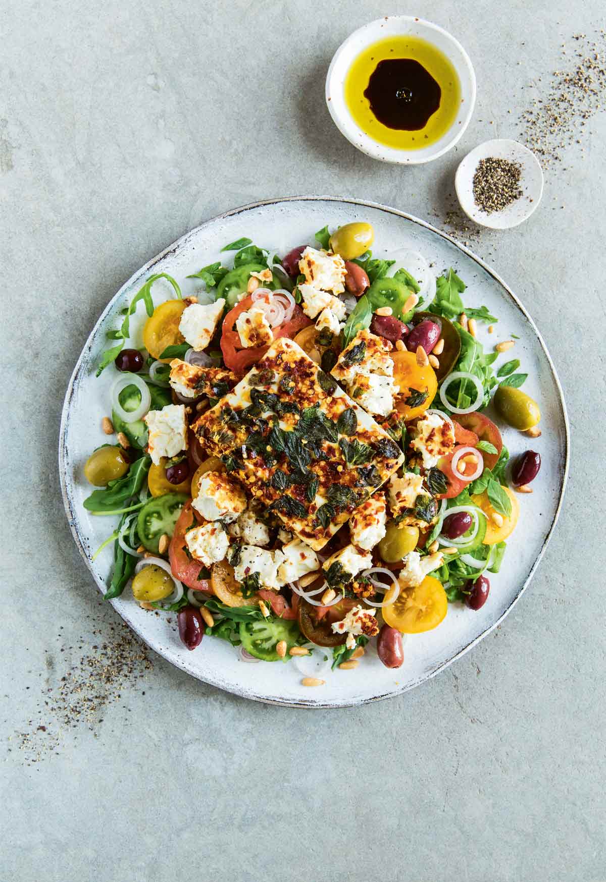 A white plate filled with tomato and feta salad with small bowls of freshly ground pepper, and olive oil and balsamic vinegar on the side.