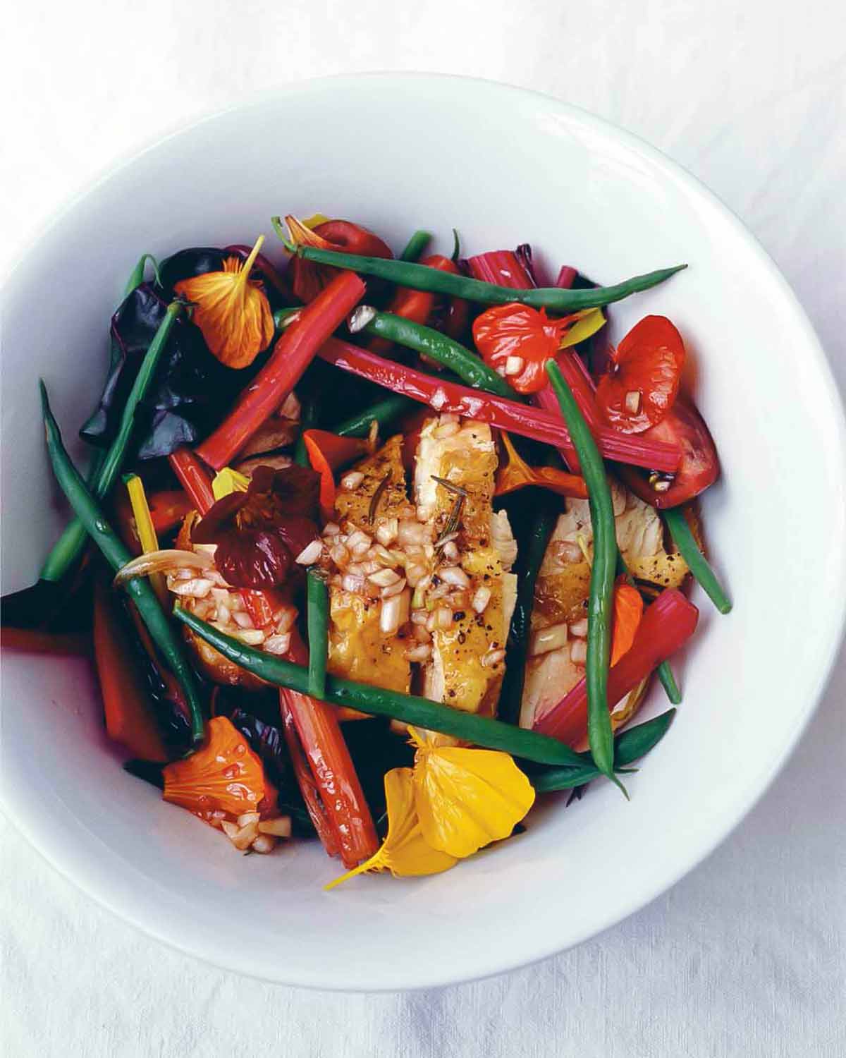 A white bowl filled with warm chicken with green beans and chard, topped with edible flowers.