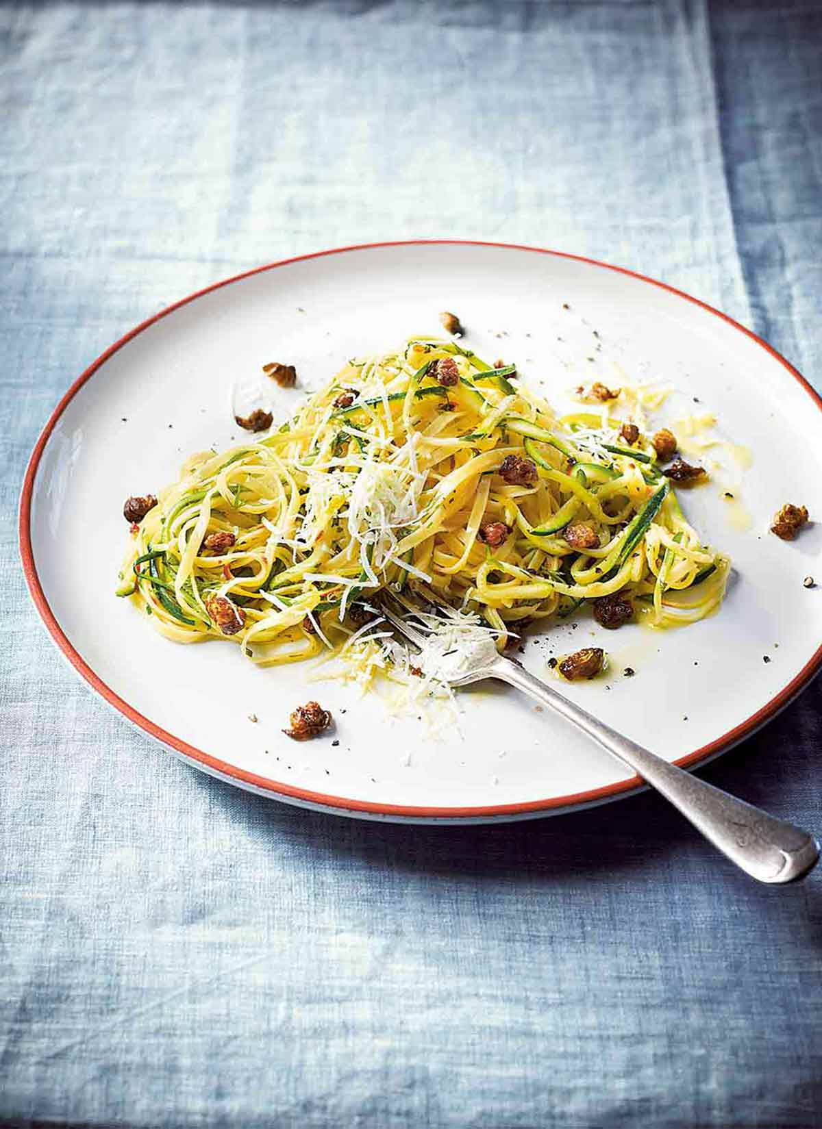 A white plate of zucchini linguine--with pasta, zucchini, garlic, capers, mint, dill, and cheese with a fork