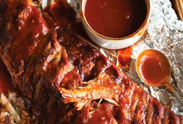 Two slabs of BBQ ribs on a piece of foil with a dish of sauce and a spoon nearby.