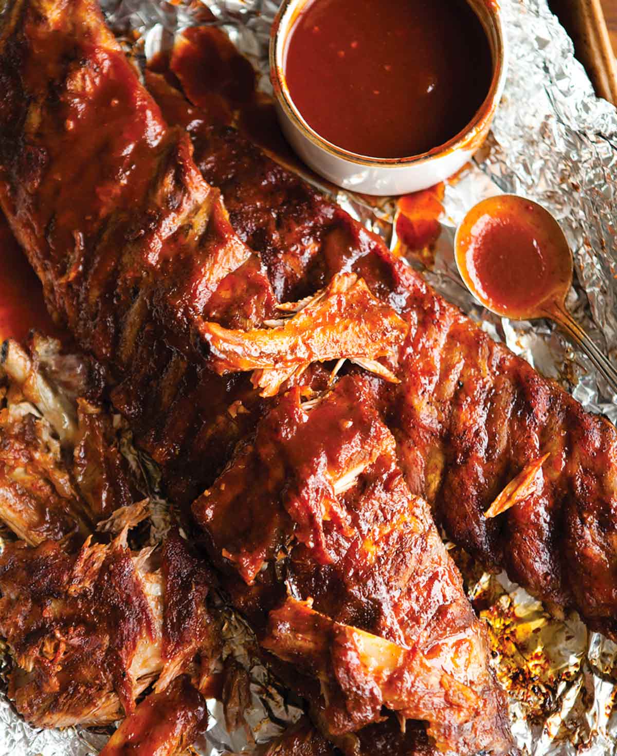 Two slabs of BBQ ribs on a piece of foil with a dish of sauce and a spoon nearby.